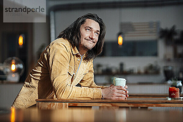 Glücklicher Mann mit Kaffeetasse lehnt am Tisch im Café