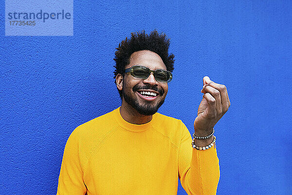 Glücklicher Mann mit Sonnenbrille gestikuliert vor blauer Wand