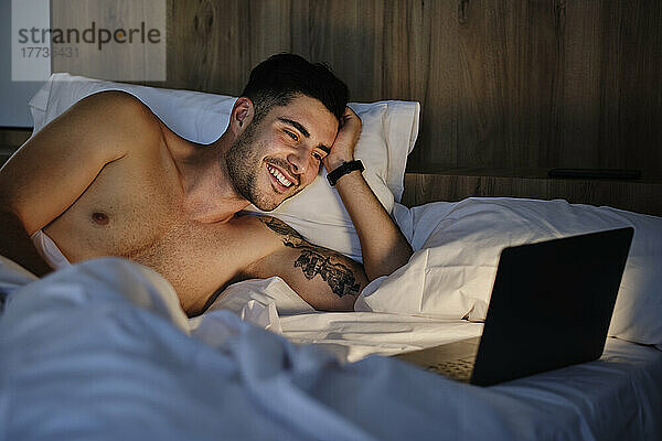 Glücklicher Mann blickt auf Laptop  der zu Hause im Bett liegt