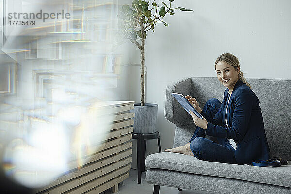 Smiling businesswoman holding tablet PC sitting on sofa in office