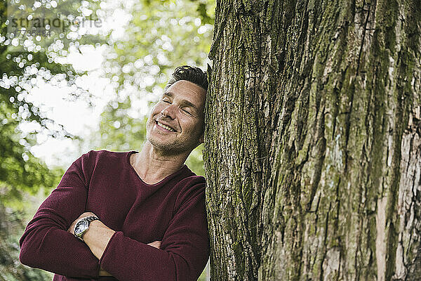 Glücklicher Mann mit geschlossenen Augen lehnt an Baum im Park