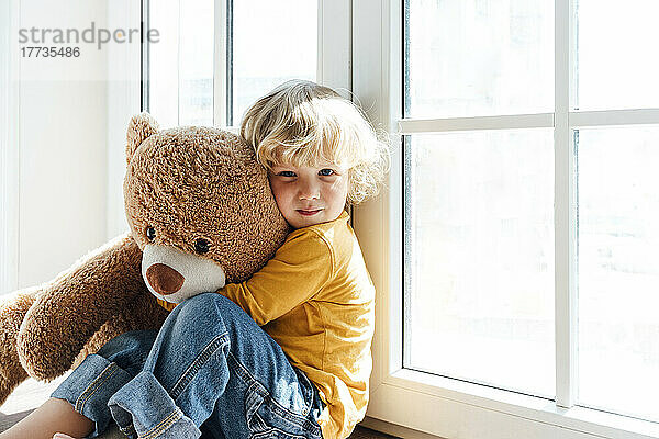 Junge mit Teddybärspielzeug sitzt zu Hause am Fenster