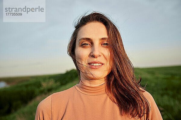 Lächelnde junge Frau mit braunen Haaren im Feld bei Sonnenuntergang