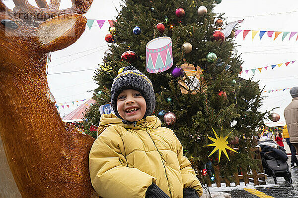 Fröhlicher Junge steht neben Hirschdekoration auf dem Weihnachtsmarkt