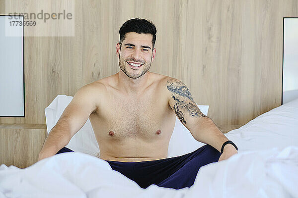Lächelnder Mann ohne Hemd  der zu Hause im Bett sitzt