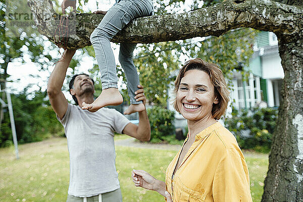 Glückliche Frau steht vor einem Mann und schaut seinen Sohn an  der auf einem Baum im Hinterhof sitzt
