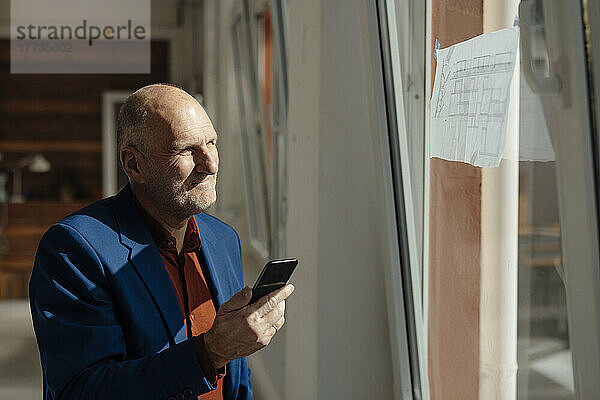 Geschäftsmann hält Mobiltelefon in der Hand und schaut auf den Bauplan am Fenster