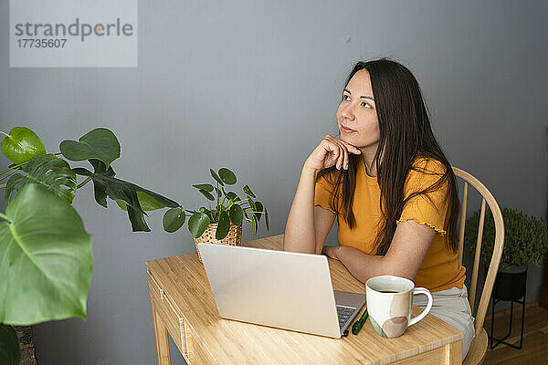 Nachdenkliche Frau sitzt mit Laptop und Kaffeetasse am Schreibtisch im Heimbüro
