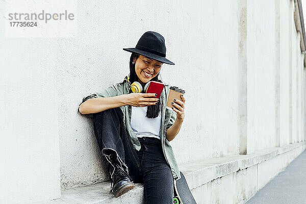 Glückliche Frau mit wiederverwendbarer Kaffeetasse und Smartphone an der Wand