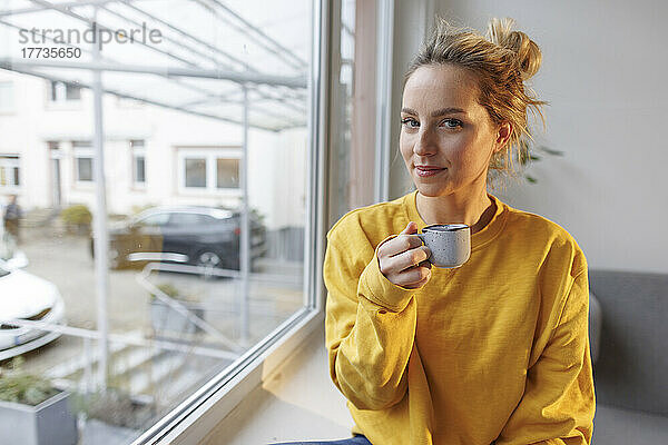 Frau hält Kaffeetasse und sitzt zu Hause auf der Fensterbank