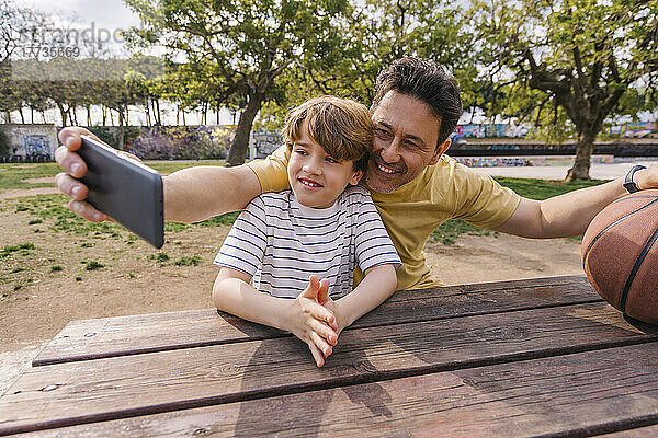 Lächelnder Mann mit Sohn  der am Tisch sitzt und ein Selfie per Smartphone macht