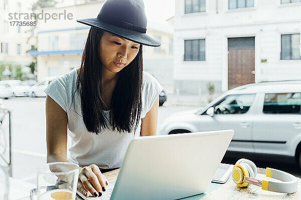Junger Freiberufler mit Hut und Laptop sitzt im Straßencafé
