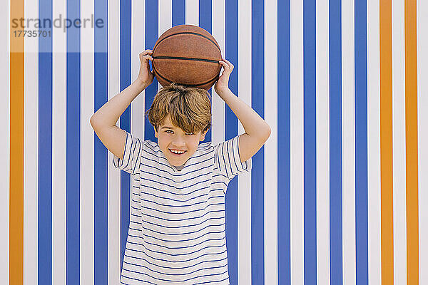 Fröhlicher Junge hält Basketball auf dem Kopf vor einer orange-blau gestreiften Wand