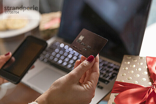 Hände einer Frau  die zu Hause Kreditkarte und Smartphone neben einem Weihnachtsgeschenk hält