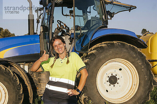 Glückliche Bäuerin gestikuliert an einem sonnigen Tag mit dem Daumen nach oben vor dem Traktor