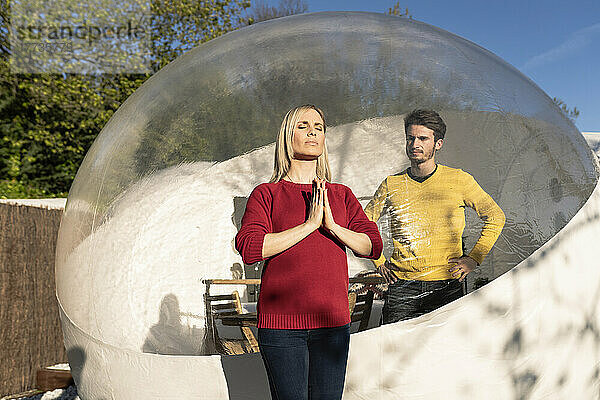 Mann in transparenter Kuppel blickt Freundin beim Yoga an einem sonnigen Tag zu