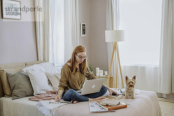 Frau schreibt in ein Buch und sitzt mit Laptop neben ihrem Hund zu Hause im Bett