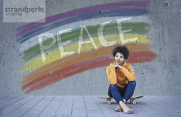 Junge Frau sitzt auf einem Skateboard vor einer Regenbogenfahne mit dem Text „Frieden“.