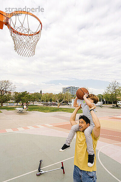 Boy with basketball sitting on father's shoulders at sports court