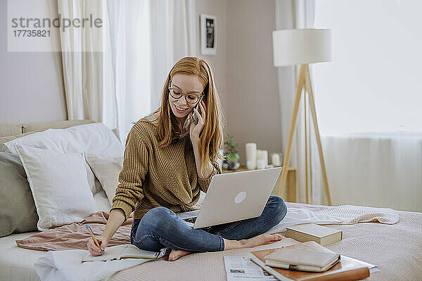 Lächelnde Frau  die mit dem Handy telefoniert und in ein Buch schreibt  sitzt mit Laptop zu Hause im Bett