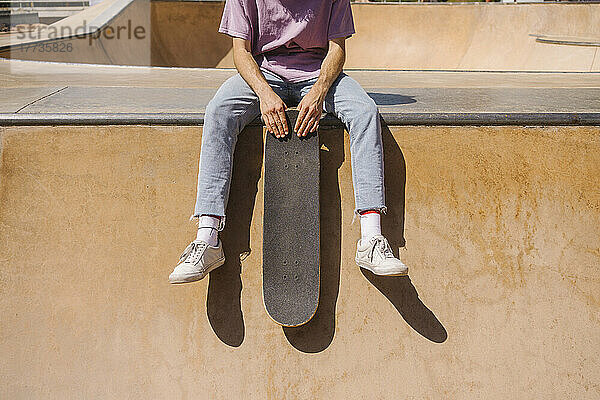 Man holding skateboard sitting at sport ramp on sunny day