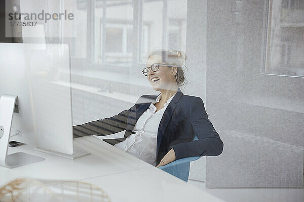 Glückliche Geschäftsfrau mit Desktop-PC am Schreibtisch  gesehen durch Glas