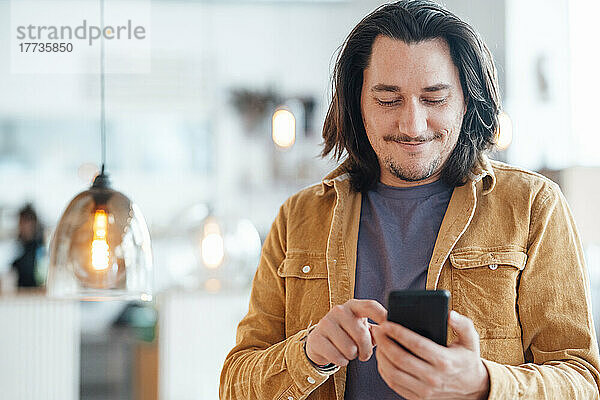 Lächelnder Hipster-Mann mit langen Haaren  der im Café sein Smartphone benutzt
