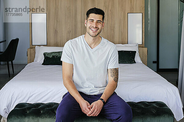 Lächelnder junger Mann  der zu Hause im Schlafzimmer sitzt