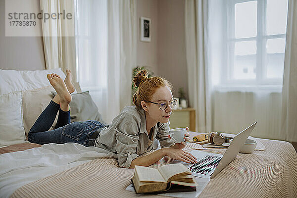 Frau mit Brille hält Kaffeetasse in der Hand und benutzt Laptop  liegt zu Hause im Bett