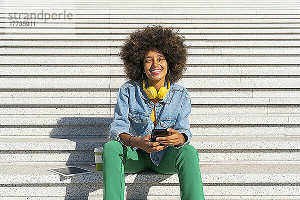 Lächelnde schöne Afro-Frau mit Smartphone sitzt neben Tablet-PC und Einweg-Kaffeetasse auf Stufen