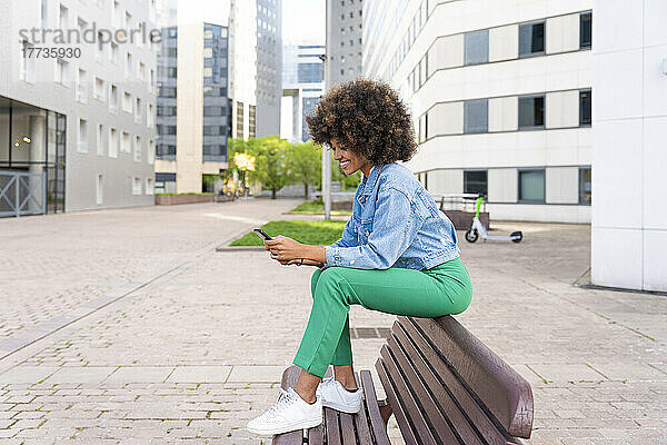 Lächelnde Afro-Frau  die SMS über ihr Smartphone sendet und auf einer Bank sitzt