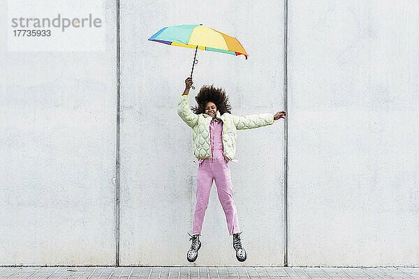 Fröhliches Mädchen springt mit Regenschirm vor einer weißen Wand