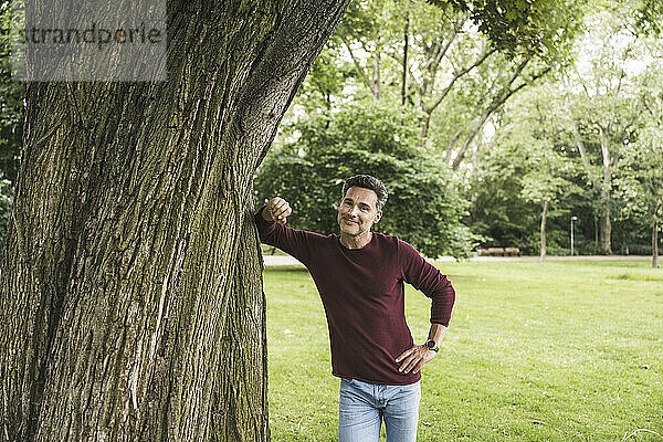 Glücklicher reifer Mann mit der Hand auf der Hüfte  der am Baum im Park steht