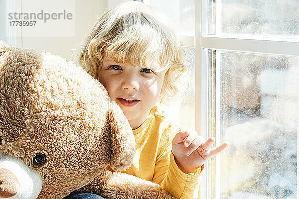 Netter Junge mit Teddybär sitzt zu Hause am Fenster