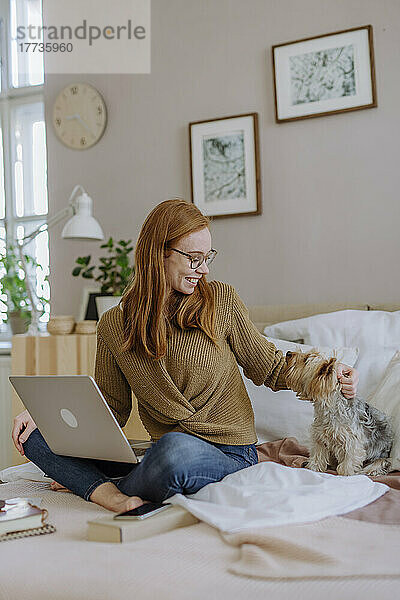 Glückliche Frau sitzt mit Laptop und streichelt ihren Hund zu Hause im Bett