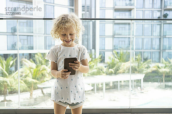 Glückliches Mädchen  das Smartphone vor dem Glasgeländer auf dem Balkon benutzt