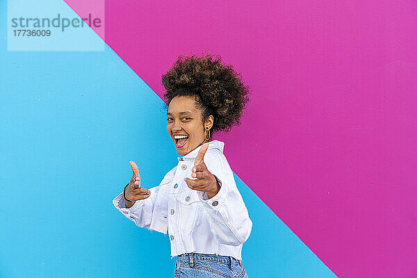 Fröhliche Frau mit Afro-Frisur gestikuliert vor rosa und blauer Wand