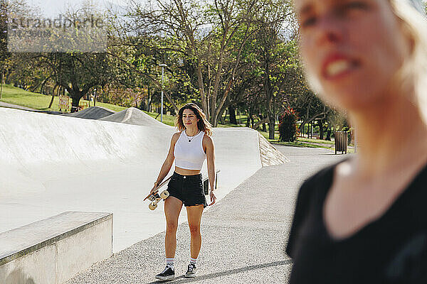 Lächelnde Frau mit Skateboard läuft an einem sonnigen Tag neben einem Freund auf der Sportrampe