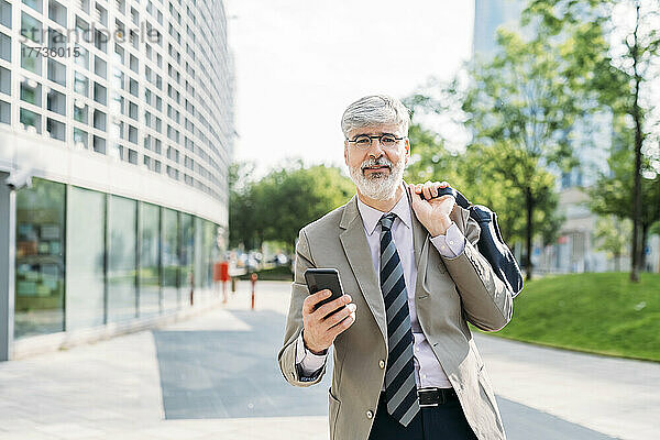 Lächelnder Geschäftsmann mit Smartphone steht vor Bürogebäude