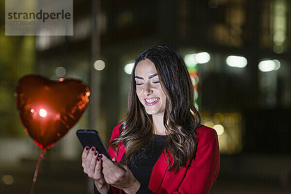 Glückliche Frau  die nachts ihr Smartphone mit einem herzförmigen Ballon benutzt