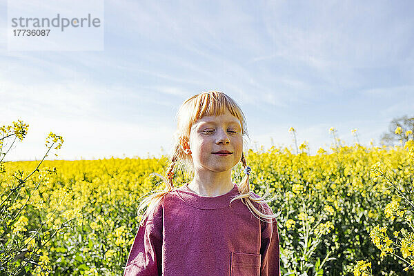 Lächelndes rothaariges Mädchen mit geschlossenen Augen steht auf einem Rapsfeld