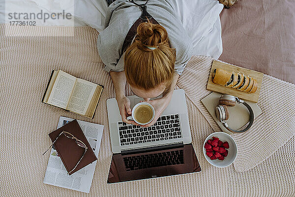 Frau mit Kaffeetasse liegt auf dem Bett und lernt zu Hause am Laptop