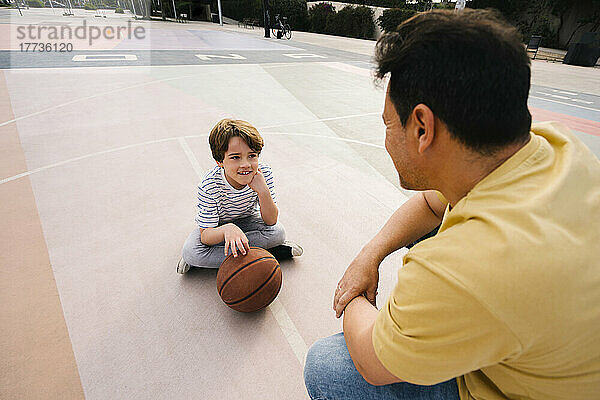Junge mit Basketball sitzt vor Vater auf Sportplatz