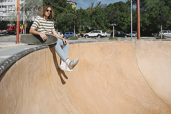 Young man with skateboard sitting at sports ramp on sunny day