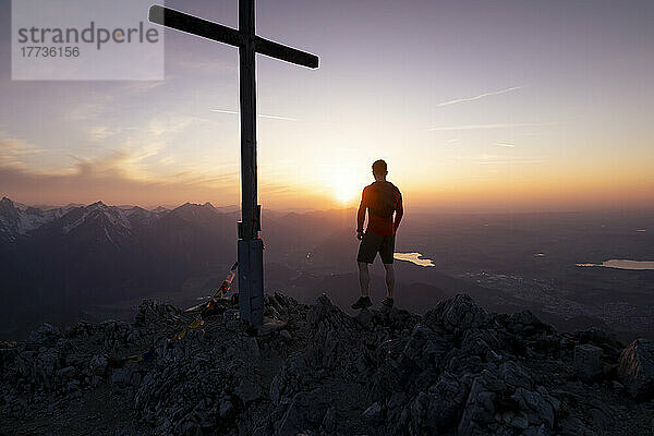 Silhouette eines Mannes  der am Gipfelkreuz auf dem Berg steht