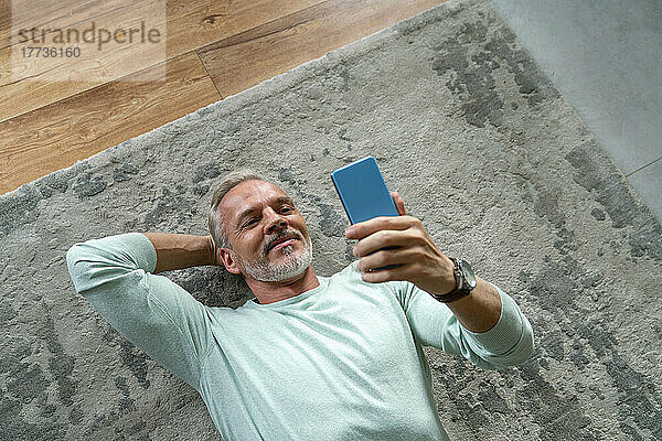 Lächelnder Mann schaut auf sein Smartphone  das auf dem Teppich liegt