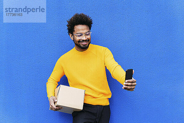 Lächelnder Lieferbote hält einen Karton in der Hand und macht ein Selfie mit dem Smartphone vor einer blauen Wand