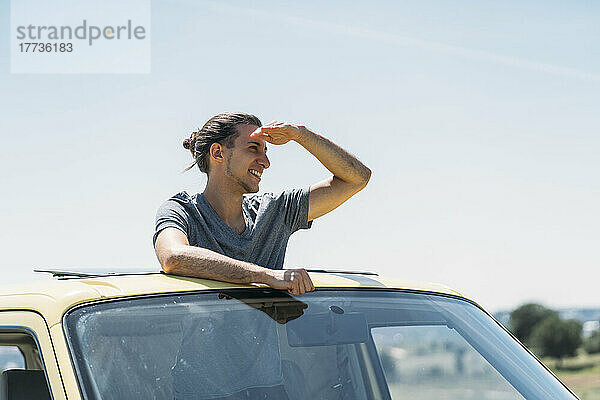 Smiling young man shielding eyes standing in van on sunny day