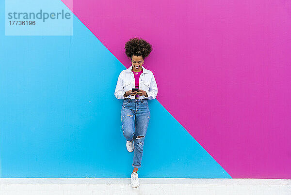 Lächelnde Frau mit Smartphone  die auf einem Bein steht und sich an eine rosa und blaue Wand lehnt