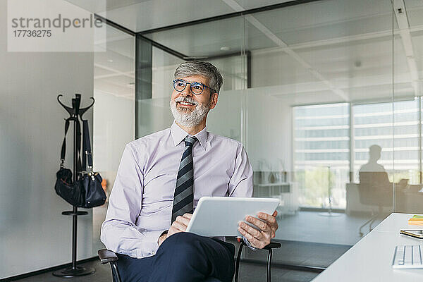 Glücklicher Geschäftsmann mit Tablet-PC  der auf einem Stuhl im Büro sitzt